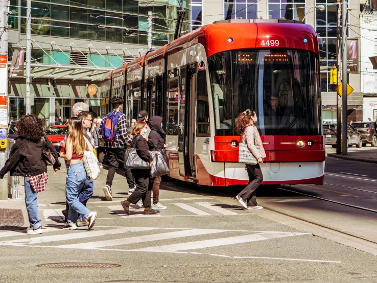 a group of people crossing a street in front of a bus, by Nicolette Macnamara, pexels contest winner, regionalism, the city of toronto, tram, 💋 💄 👠 👗, bright sunny day