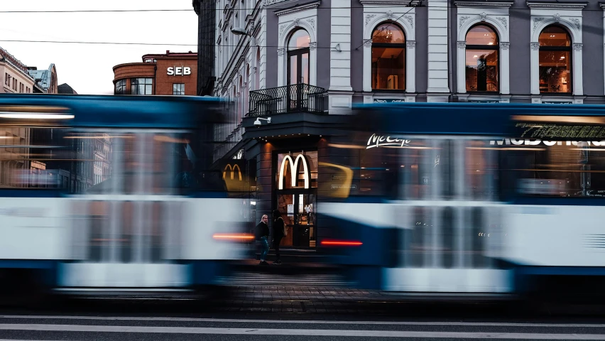 a blue and white bus driving down a street next to a tall building, by Micha Klein, pexels contest winner, hyperrealism, mcdonalds restaurant, tram, motion graphic, finland