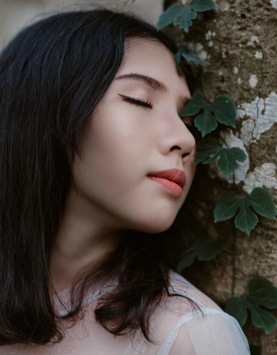 a woman leaning against a tree with her eyes closed, inspired by Yu Zhiding, trending on pexels, aestheticism, with ivy, south east asian with round face, iridescent skin, prominent cheekbones