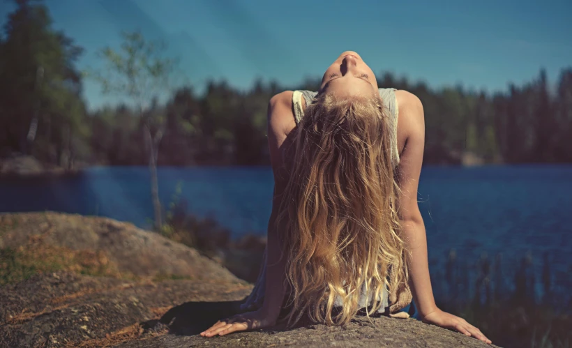 a woman sitting on top of a rock next to a lake, inspired by Elsa Bleda, unsplash, happening, blonde flowing hair, in the sun, head tilted down, retro stylised