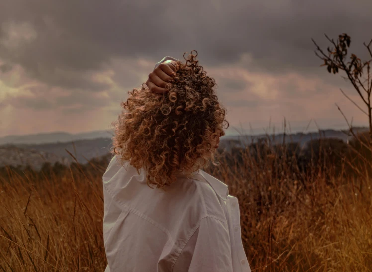 a woman with curly hair standing in a field, trending on pexels, her hair is tied above her head, gloomy skies, in the hillside, her hair is white