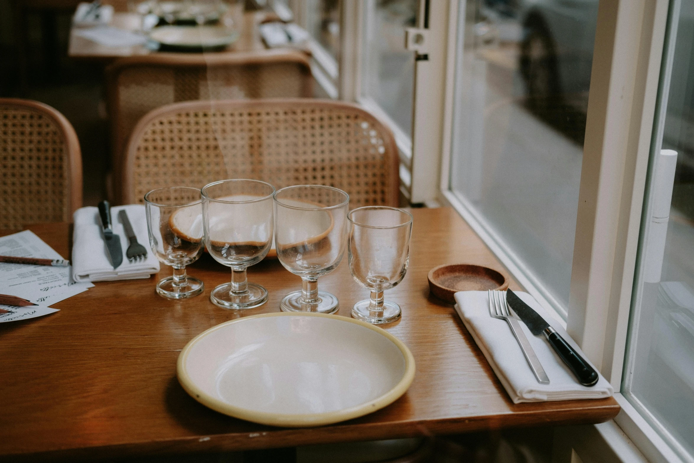 a wooden table topped with a white plate next to a window, unsplash, french, american canteen, glassware, sydney hanson
