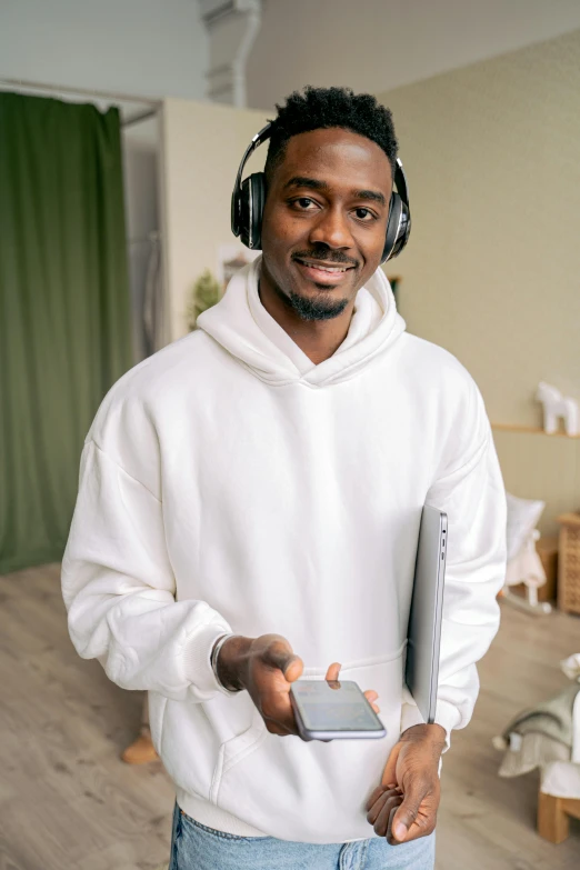 a man standing in a living room with headphones on, wearing a white sweater, man is with black skin, tech robes, holding notebook