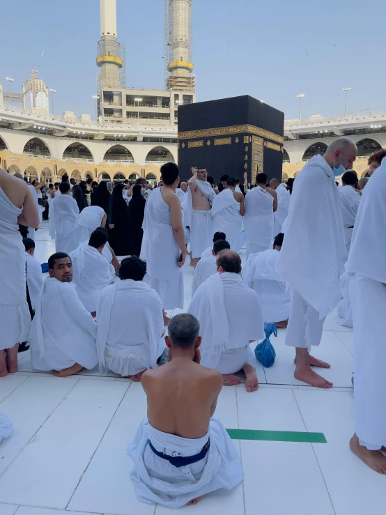 a group of people sitting on the ground in front of a building, golden-white robes, mekka, wearing nothing, reddit post