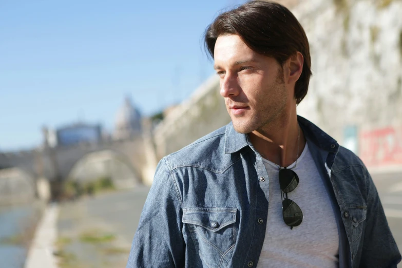 a man standing next to a body of water, a portrait, inspired by Romano Vio, shutterstock, wearing double denim, rome in background, with dark brown sunglasses, antony starr