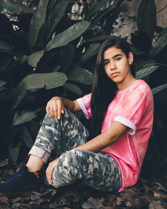 a woman sitting on the ground in front of a plant, by Isabel Naftel, trending on unsplash, wearing a tie-dye t-shirt, portrait androgynous girl, estefania villegas burgos, wearing camo