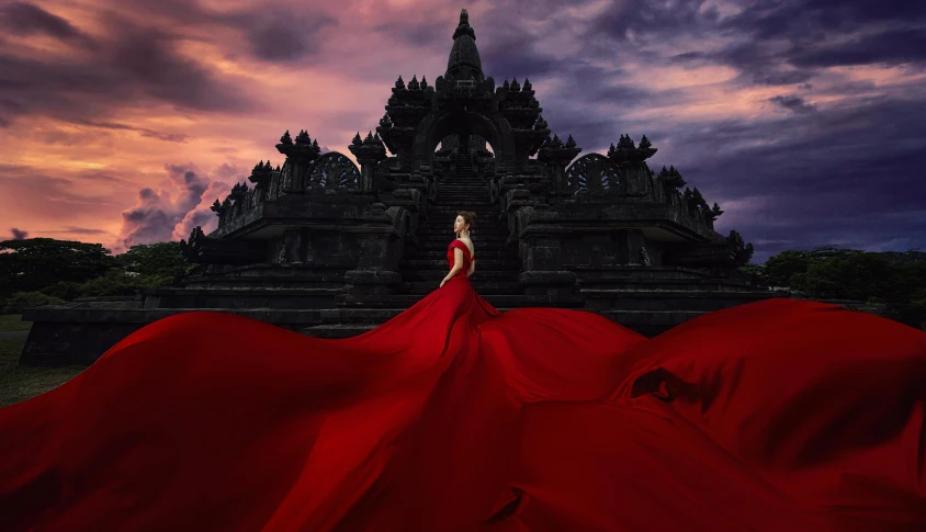 a woman in a red dress standing in front of a building, an album cover, inspired by Rudy Siswanto, pexels contest winner, art photography, at borobudur, swirling red-colored silk fabric, gothic regal, rendering of beauty pageant