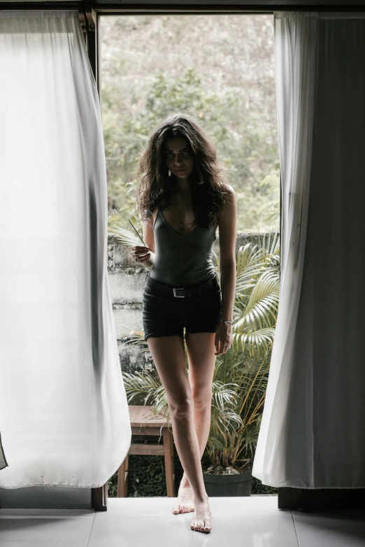 a woman standing in front of a window, inspired by Nan Goldin, pexels contest winner, renaissance, sexy girl wearing shorts, long dark curly hair, bali, full body! pretty face