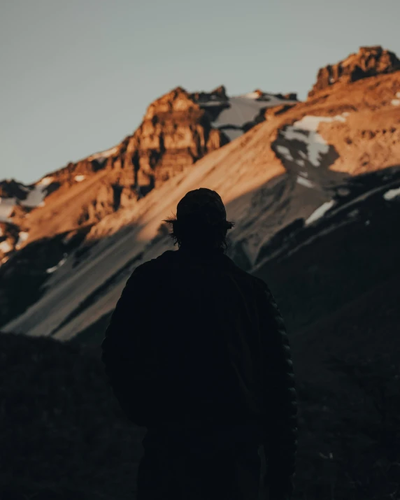 a person standing in front of a mountain, back lit, snow capped mountains, looking sad, trending photo