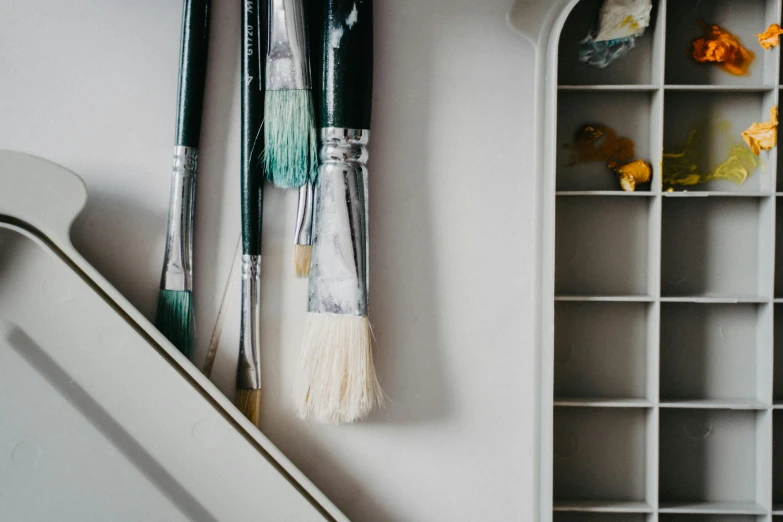 a bunch of paint brushes sitting on top of a shelf, inspired by Kyffin Williams, trending on pexels, arts and crafts movement, white marble interior photograph, white broom closet, white and teal metallic accents, stands at a his easel