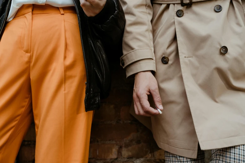 a couple of people standing next to each other, trending on pexels, trench coat, white and orange, lesbians, brown pants