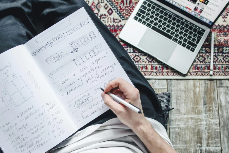 a man sitting on top of a wooden floor next to a laptop computer, a sketch, by Julia Pishtar, pexels contest winner, schematic in a notebook, 9 9 designs, writing in journal, thumbnail