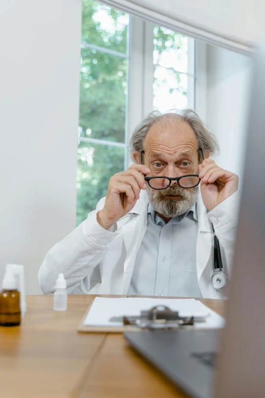 a man sitting at a desk in front of a computer, a picture, shutterstock, hyperrealism, wearing lab coat and glasses, he is a mad old man, doctors mirror, wearing a grey robe