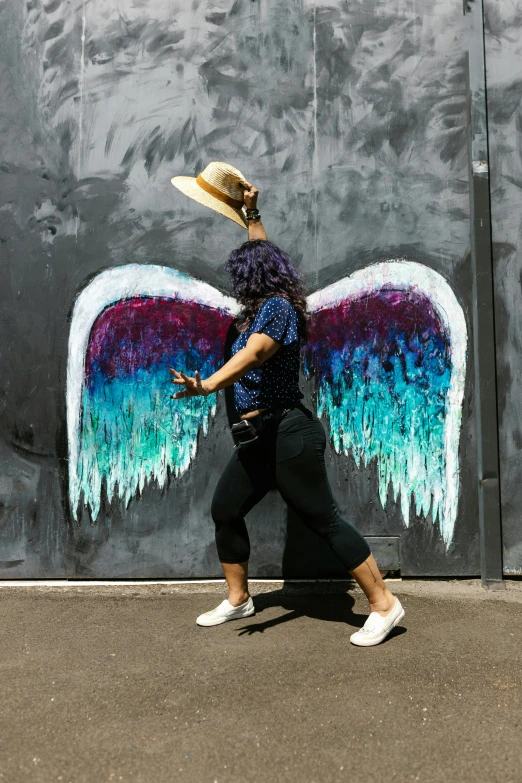 a woman walking in front of a wall with wings painted on it, chalk art, by Meredith Dillman, pexels contest winner, street art, black and blue and purple scheme, bum, angelheaded hipsters, te pae