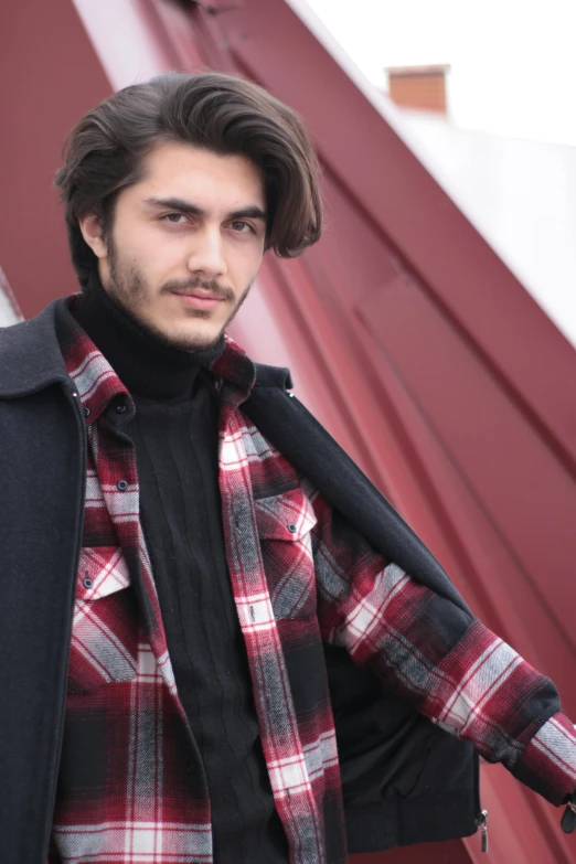 a man with long hair and plaid shirt is standing near a red wall