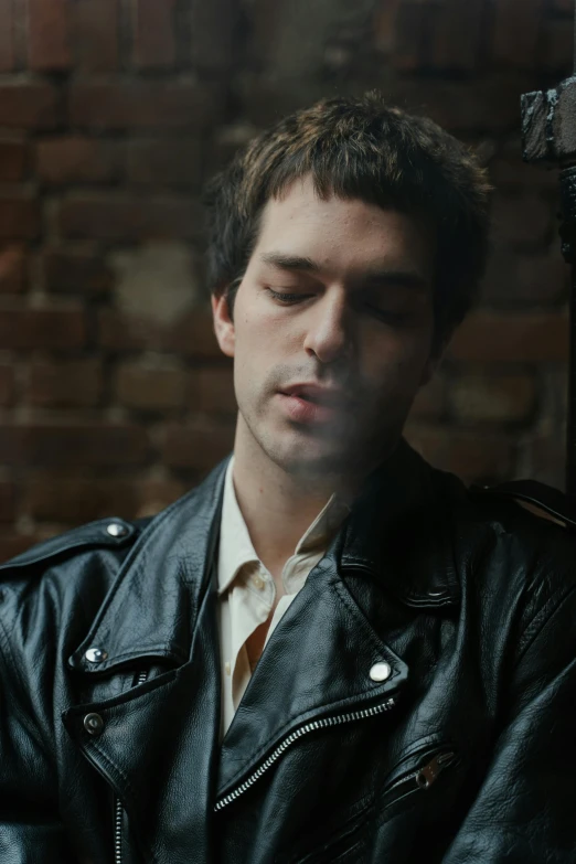 a man in a black leather jacket sits next to a brick wall