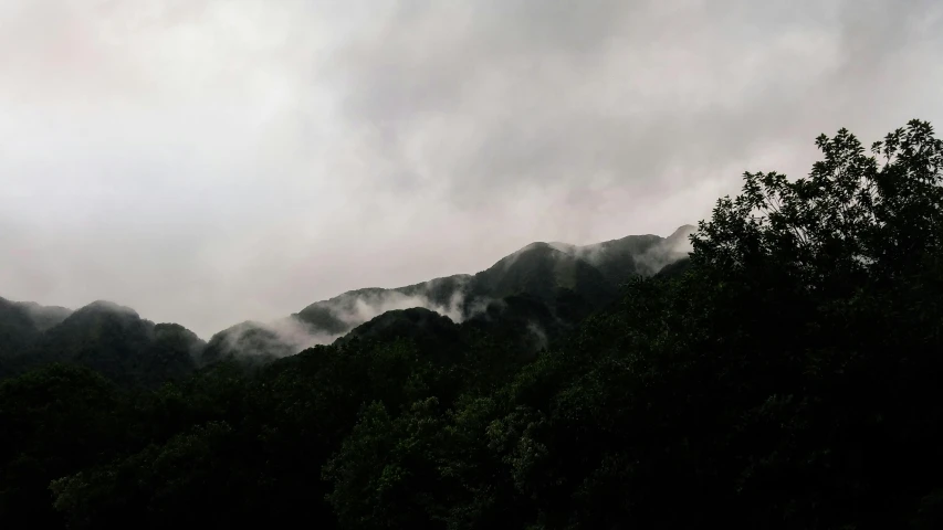 a mountain covered in clouds with trees in the foreground, inspired by Sesshū Tōyō, unsplash, hurufiyya, low quality photo, rainy and gloomy atmosphere, low angle photograph, rinko kawauchi