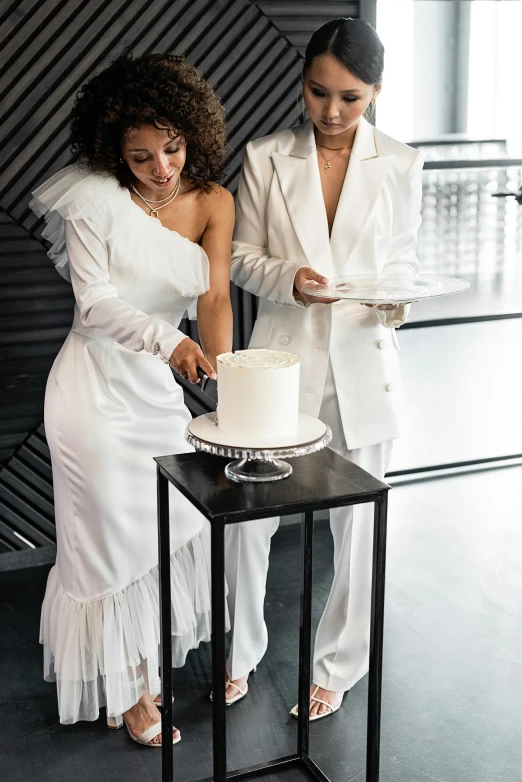 a couple of women standing next to a cake, white suit, sleek robes, white table, celebrating