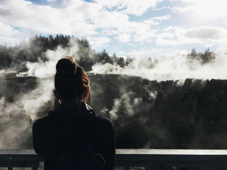 a woman standing on top of a bridge next to a river, geysers of steam, looking outside, te pae, instagram post