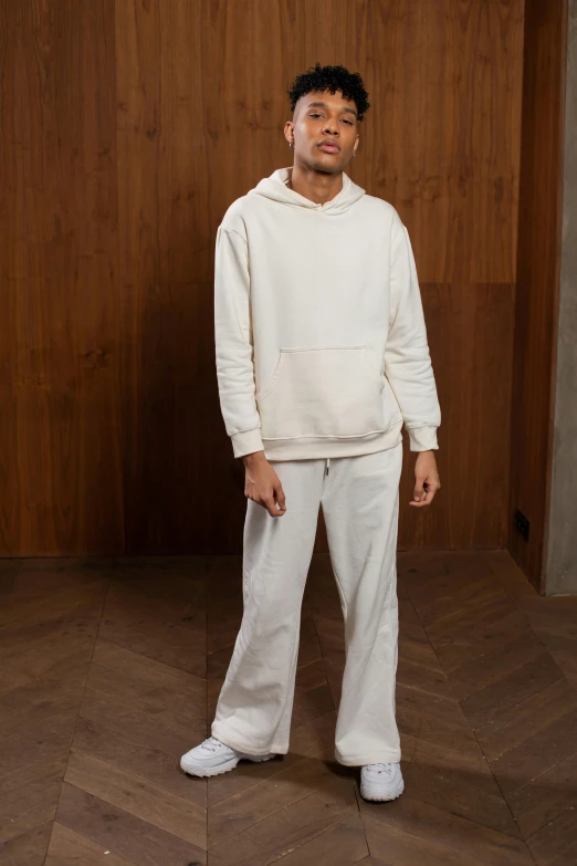 a man standing in front of a wooden wall, inspired by Raphaël Collin, wearing a hoodie and sweatpants, wearing white pajamas, official store photo, offwhite