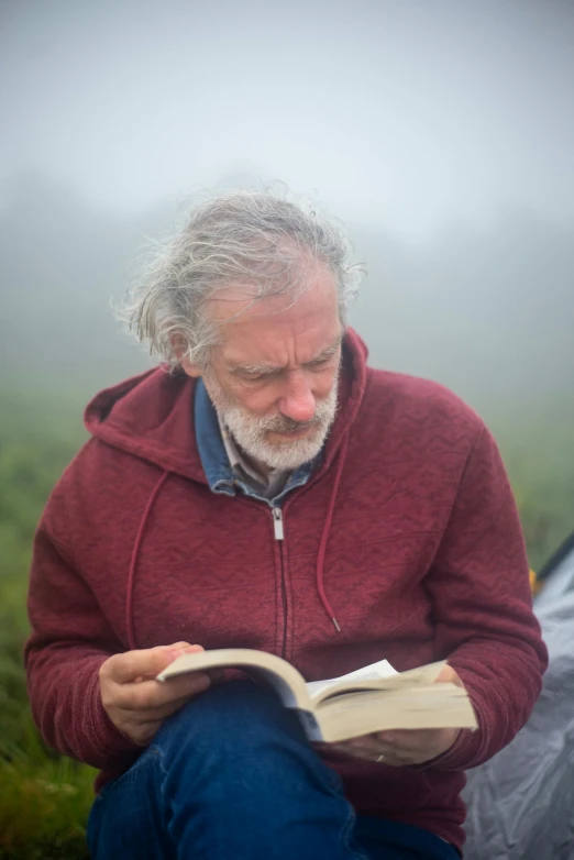 a man sitting in front of a tent reading a book, by Bedwyr Williams, some grey hair in beard, under a gray foggy sky, pastoral, 2019 trending photo