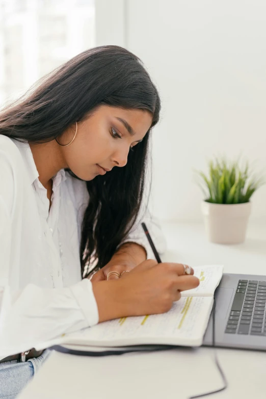 a woman sitting at a desk working on a laptop, pexels contest winner, academic art, young woman with long dark hair, writing on a clipboard, south east asian with long, white hue