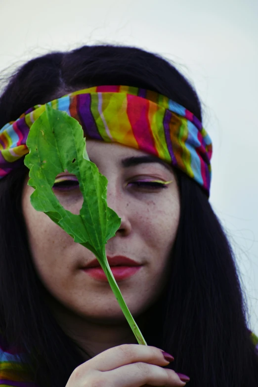 a woman holding a leaf in front of her face, an album cover, unsplash, rojava, blindfold, heterochromia, summer of love