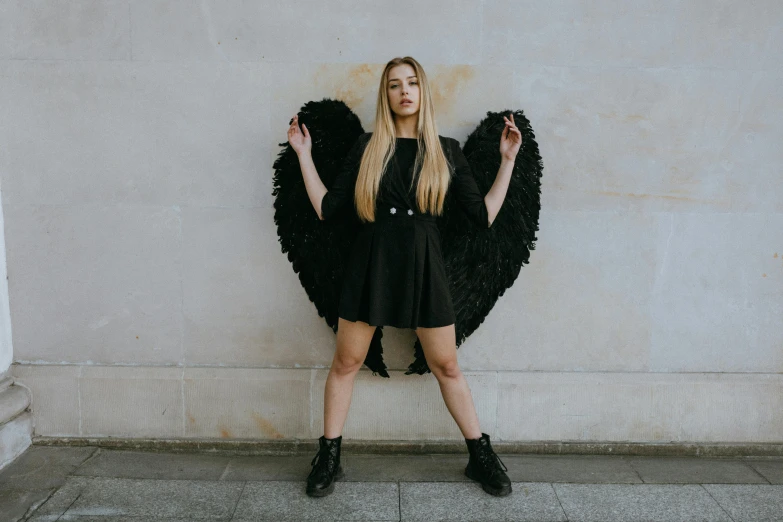 a woman in a black dress standing in front of a wall, an album cover, pexels contest winner, international gothic, winged girl angel, sydney sweeney, full body with costume, cosplay photo