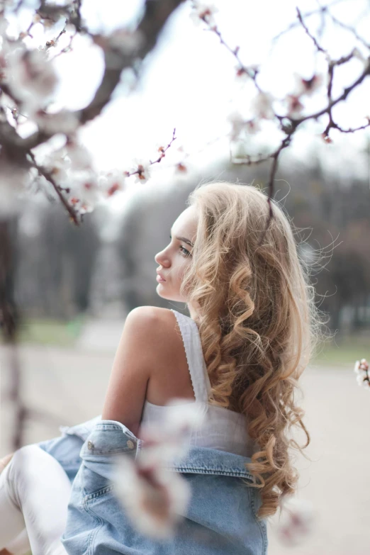 a woman sitting on a bench in front of a tree, a picture, trending on unsplash, renaissance, long fluffy curly blond hair, blossoms, ukrainian girl, over the shoulder view