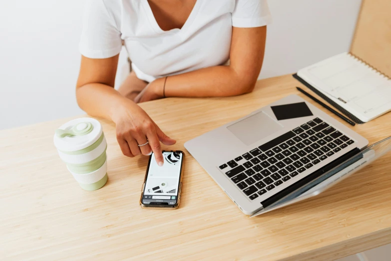 a woman sitting at a table with a laptop and a cell phone, by Carey Morris, trending on pexels, worksafe. instagram photo, plain background, scrolling computer mouse, 9 9 designs