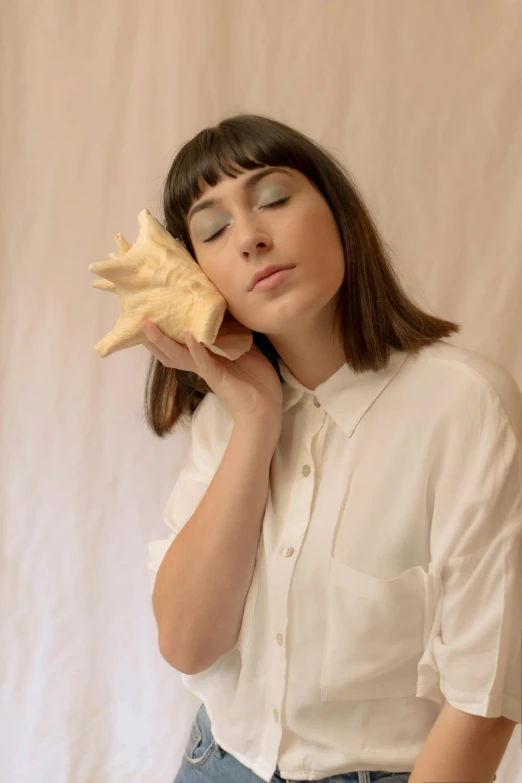 a woman holding a seashell up to her face, an album cover, by Ellen Gallagher, trending on pexels, magic realism, organic ceramic white, shirt, ana de la reguera portrait, from side