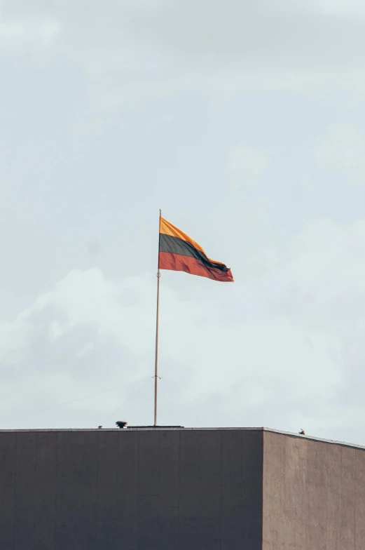 a flag on top of a building on a cloudy day, by Attila Meszlenyi, unsplash, berlin secession, yellows and reddish black, sri lanka, black and orange, square