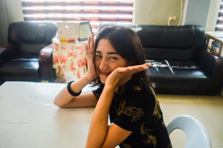 a woman sitting at a table with her hands on her face, a picture, inspired by Yuko Tatsushima, pexels contest winner, waving at the camera, malaysian, profile image, sitting on the floor