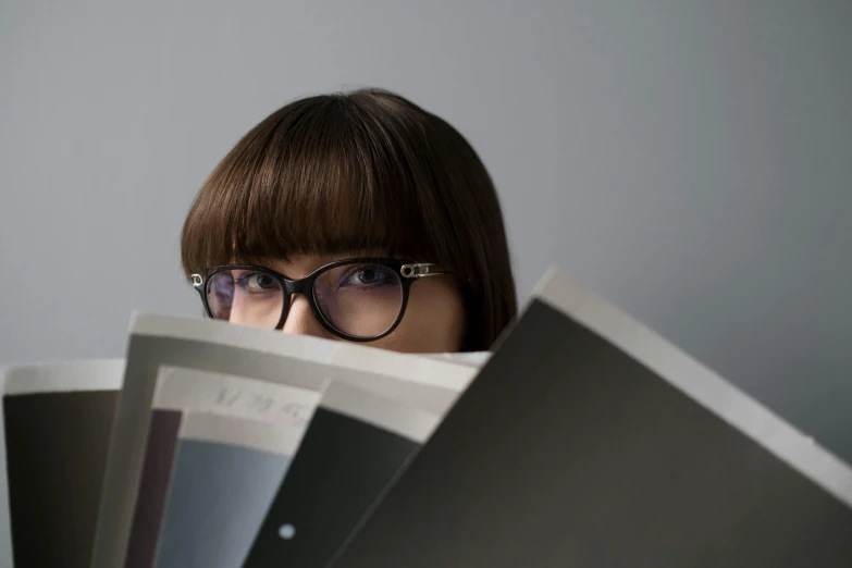 a woman wearing glasses and reading a book, trending on unsplash, private press, on grey background, hiding, magazines, looking up at camera