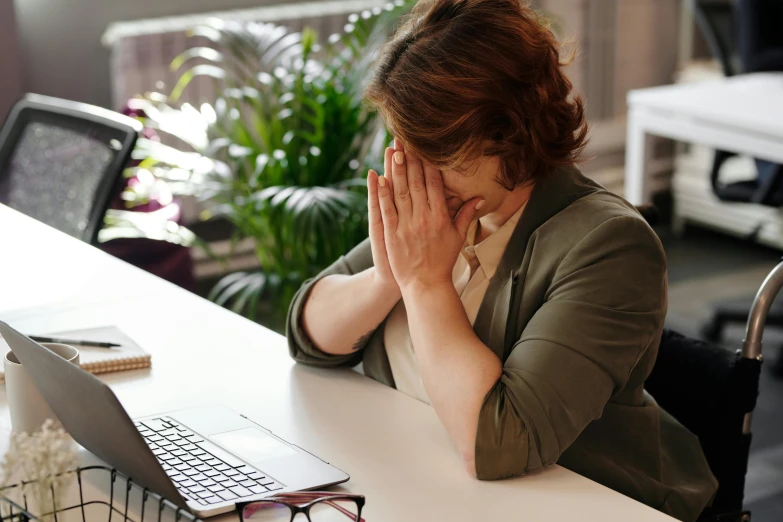 a woman sitting at a desk with her hands on her face, trending on pexels, worksafe. instagram photo, devastated, bent - over posture, non-binary