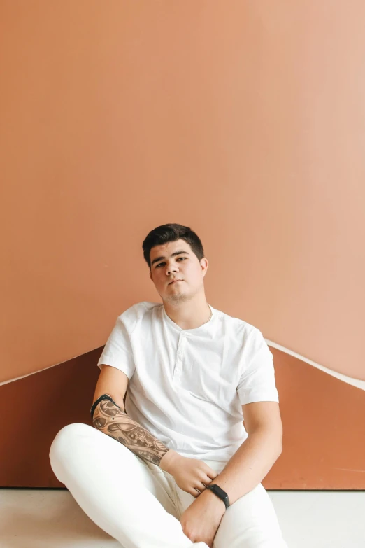 a man sitting on the ground in front of a wall, an album cover, by Jessie Alexandra Dick, trending on pexels, man in white t - shirt, sitting on a mocha-colored table, portrait of archie andrews, confident looking