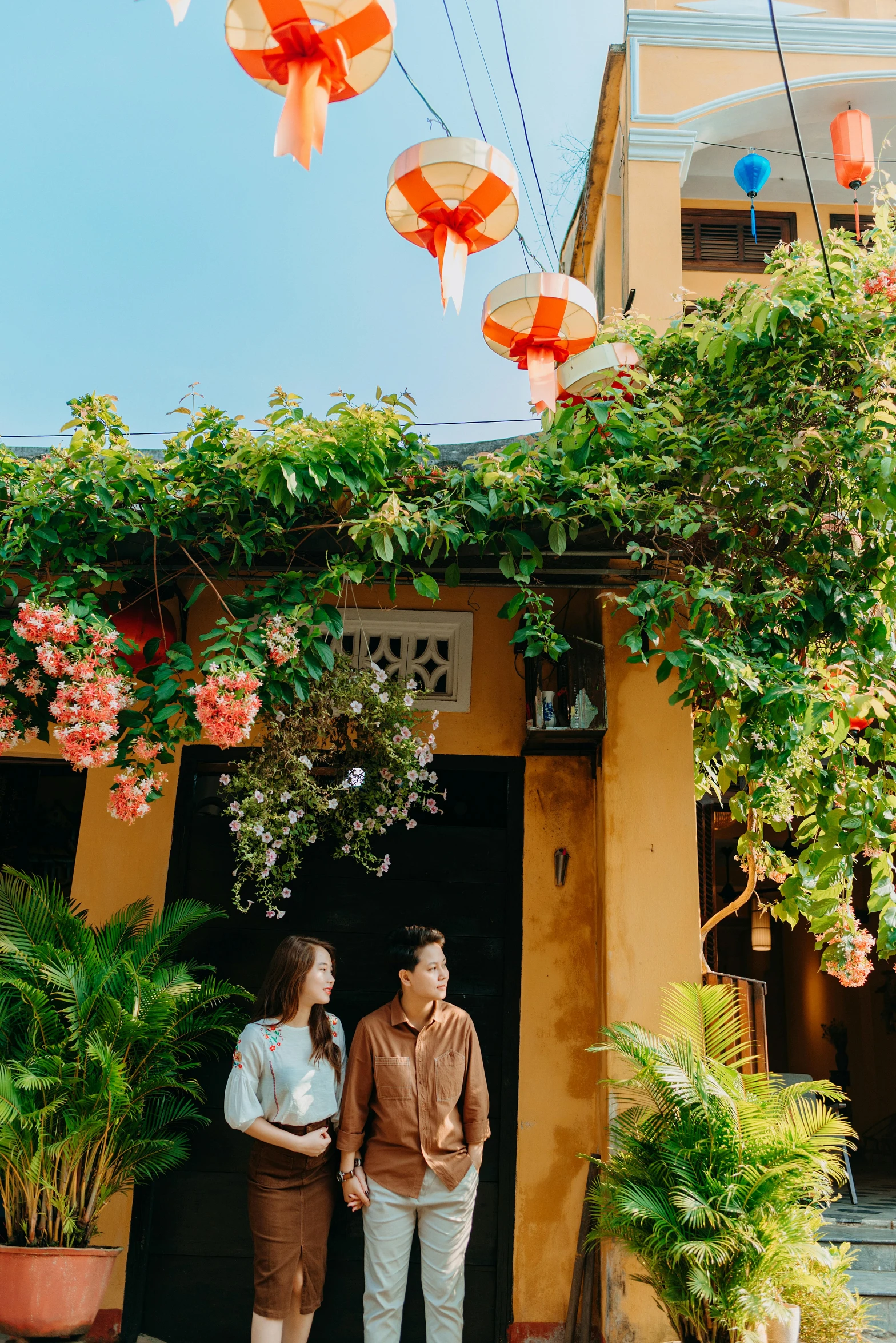 a man and a woman standing in front of a building, lush plants and lanterns, in style of lam manh, exterior, flowers and vines