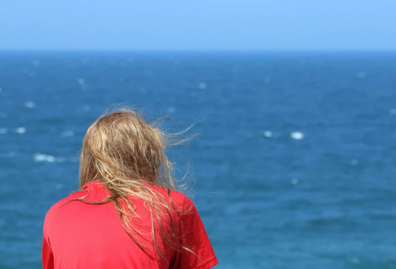 a woman in a red shirt looking out at the ocean, pexels contest winner, a blond, aged 13, view from far away, blue and red