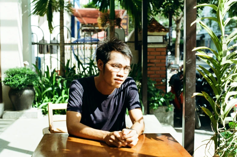 a man sitting at a wooden table outside, a portrait, by Julia Pishtar, unsplash, shin hanga, malaysian, in a bright cafe, avatar image, around 1 9 years old