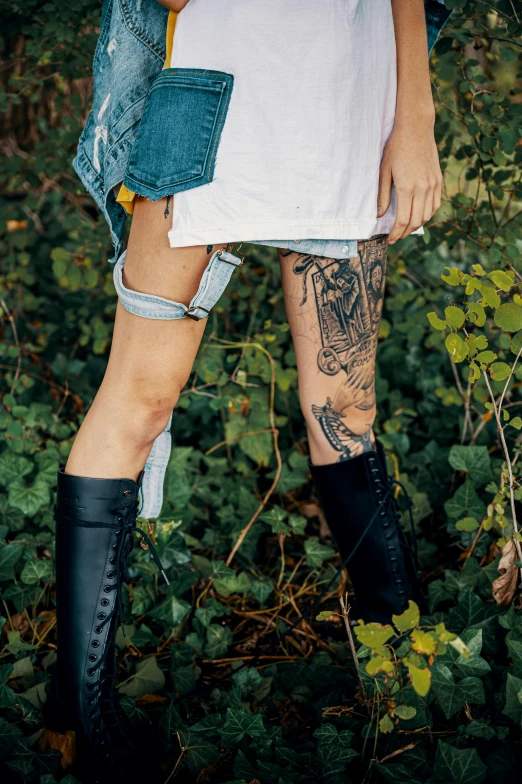 a woman that is standing in the grass, a tattoo, trending on pexels, riding boots, thigh skin, lush greens, wearing punk clothing