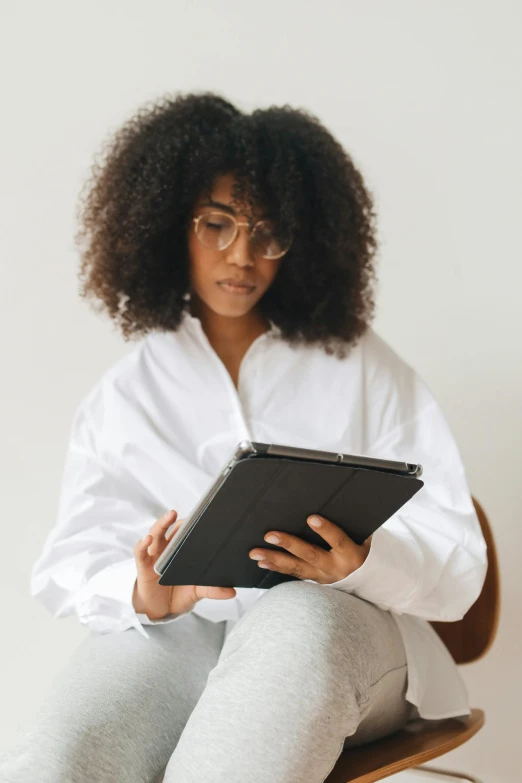 a woman sitting on a chair holding a tablet, by Carey Morris, trending on pexels, afrofuturism, white dress shirt, long afro hair, reading glasses, wearing a grey robe