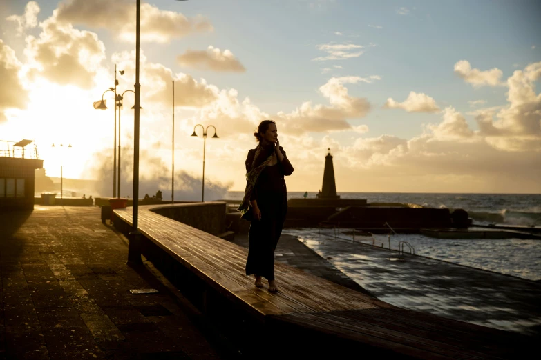 a woman standing on a pier next to the ocean, by Jesper Knudsen, pexels contest winner, happening, late afternoon sun, azores, walking to work, cloak flittering in the wind