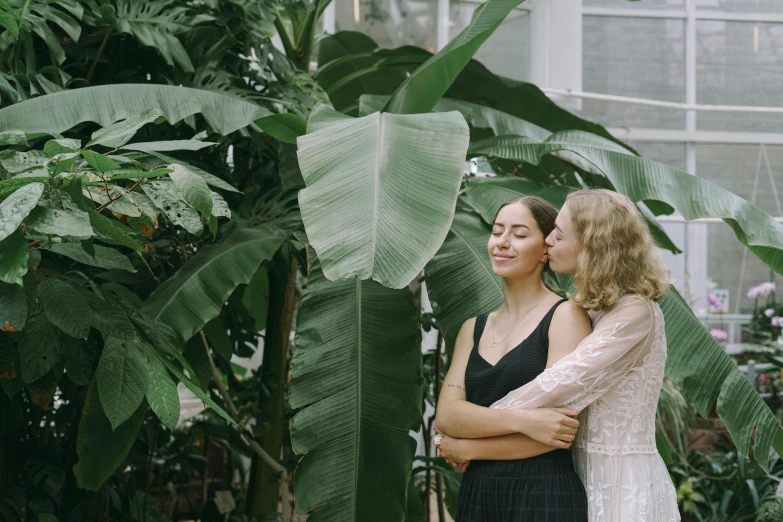 a couple of women standing next to each other, by Emma Andijewska, pexels contest winner, romantic greenery, biodome, lesbian embrace, monstera deliciosa