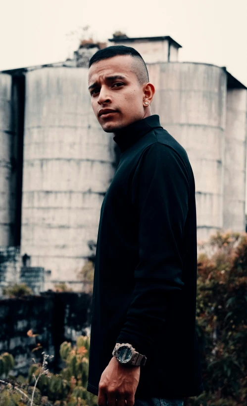 a man in a black shirt standing in front of a silos, an album cover, inspired by Byron Galvez, pexels contest winner, wearing a turtleneck and jacket, shaved sides, profile image, looking towards camera