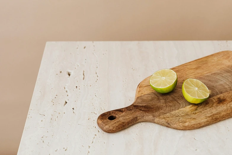 a wooden cutting board with two limes on it, a still life, inspired by Gordon Browne, trending on pexels, minimalism, white travertine terraces, beige cream natural muted tones, background image, at the counter