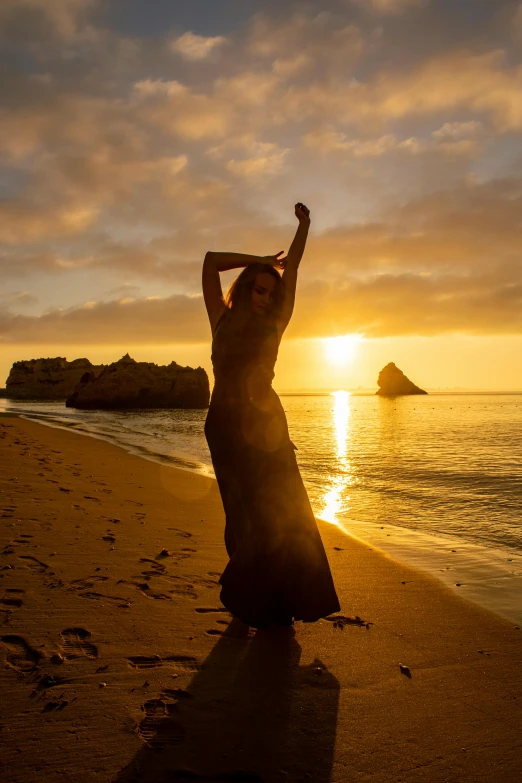 a woman wearing a long dress on the beach as the sun sets