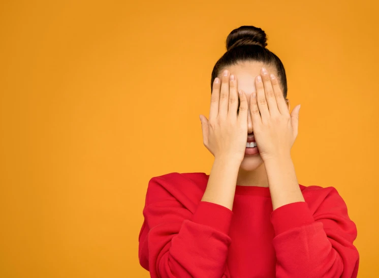 a woman covering her face with her hands, trending on pexels, pop art, red and orange color scheme, mischievous look, shoulder eyes, yellow backdrop