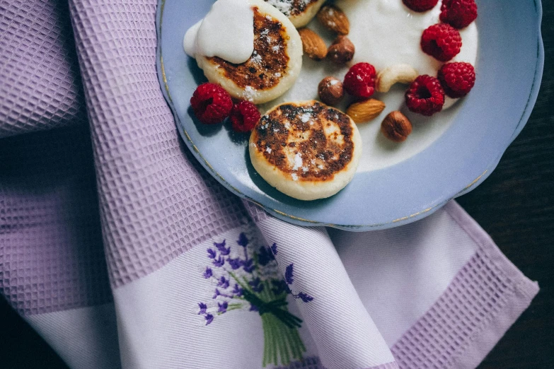 a blue plate topped with pancakes and raspberries, inspired by Ödön Márffy, unsplash, romanticism, cozy calm! fabrics textiles, white and purple, background image, photograph 3 5 mm