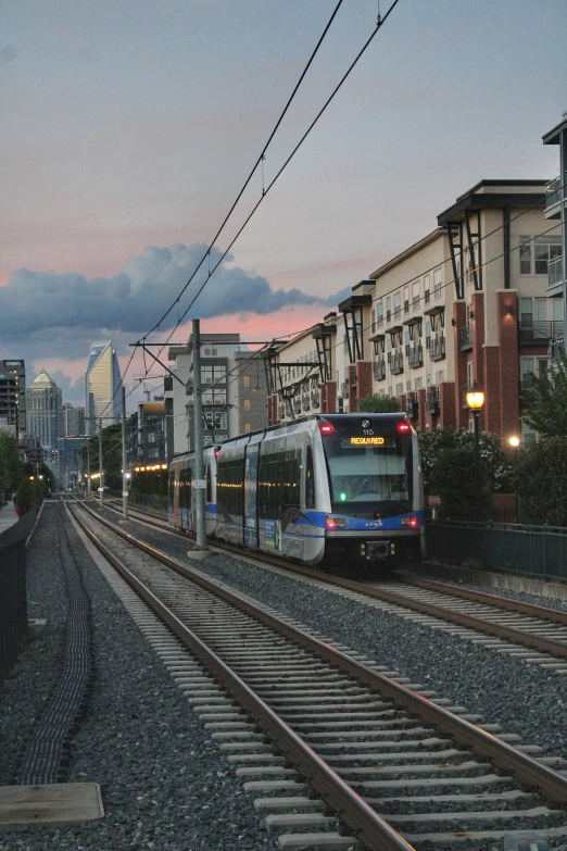 a train traveling down train tracks next to tall buildings, at dusk, ballard, orange line, square