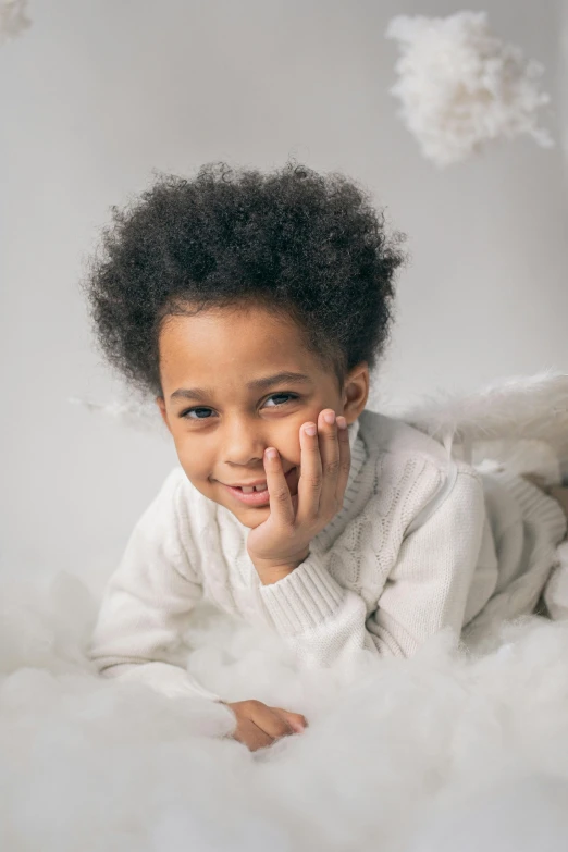 a little girl laying on top of a fluffy white blanket, inspired by Louis Le Nain, with afro, winged boy, light grey backdrop, while smiling for a photograph